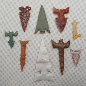 May include: A collection of nine antique arrowheads in various shapes and colors. The arrowheads are made of stone and feature shades of brown, green, red, and white. The collection includes triangular and stemmed designs, showcasing ancient craftsmanship.