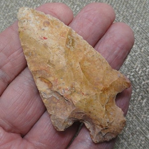 May include: A tan and light brown stone arrowhead with a pointed tip and a notched base. The surface of the arrowhead features a textured, mottled appearance with visible flaking patterns and subtle variations in color throughout the stone.