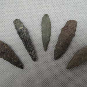 May include: Five ancient stone arrowheads of varying sizes and colors are arranged on a white surface. The arrowheads range in color from gray to brown, with a rough, textured appearance. The central arrowhead is a pale green color.