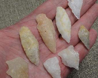 May include: A collection of eight small, pale-colored stone arrowheads and fragments. The arrowheads vary in shape, with some pointed and others triangular. The stones are held in a hand, showcasing their natural textures and hues.