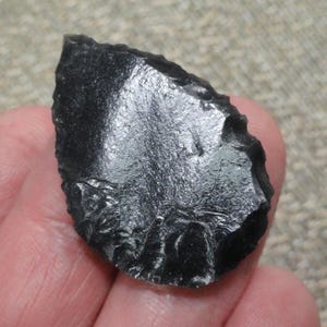 May include: A black obsidian arrowhead or tool with a rough, chipped edge. The surface of the obsidian is smooth and reflective, catching the light. The shape is roughly triangular, with a pointed tip.