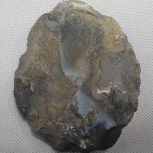 May include: A roughly oval-shaped, gray and brown stone tool with a chipped edge. The stone has areas of lighter gray and tan, with a smooth, polished appearance in some areas. The tool appears to be a prehistoric hand axe or scraper.