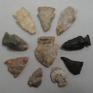 May include: A collection of nine stone arrowheads arranged in a circular pattern. The arrowheads vary in color from cream, tan, and grey to black. The central arrowhead features black text that reads MERCER CO. OH. ST. MARYS O.