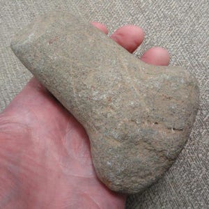 May include: A gray stone tool with a rounded handle and a rectangular head. The stone has a rough, textured surface and appears to be hand-shaped. The tool is held in a person's hand.
