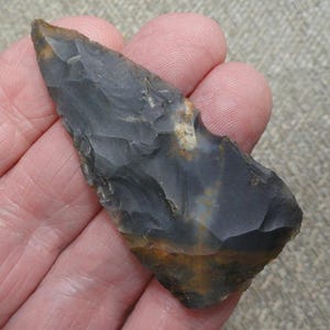 May include: A hand-held, dark gray flint arrowhead with a sharp point and a rough, textured surface. The arrowhead has brown and orange hues near the base. The artifact is likely from an ancient civilization.