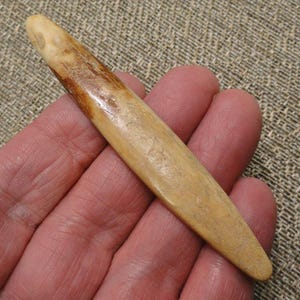 May include: A smooth, elongated, and tapered object with a light tan and brown mottled surface. The item features a rounded, pointed end and a slightly wider, rounded base, displaying a natural, polished texture with visible grain-like markings.