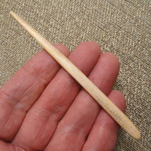 May include: A slender, tapered, off-white wooden needle or stylus with a smooth, polished surface and a pointed tip. The object features a light beige color with subtle, natural grain patterns running along its elongated, cylindrical body.