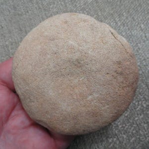 May include: A circular, tan-colored stone with a rough, textured surface. The object has a flattened, disc-like shape with visible natural variations in its earthy tone and a slightly irregular, rounded edge along its perimeter.