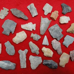 May include: A collection of various stone arrowheads and tools in shades of gray, tan, and pink, arranged on a red background. The artifacts vary in size and shape, showcasing different designs and textures.