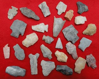 May include: A collection of various stone arrowheads and tools in shades of gray, tan, and pink, arranged on a red background. The artifacts vary in size and shape, showcasing different designs and textures.