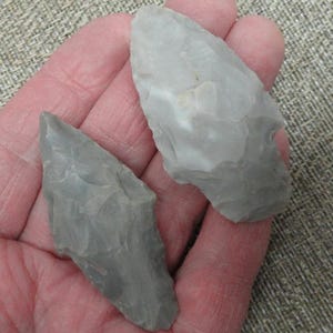May include: Two gray, hand-knapped stone arrowheads. The arrowheads are held in a hand, showcasing their sharp, pointed tips and textured surfaces. The stone has a slightly translucent quality.