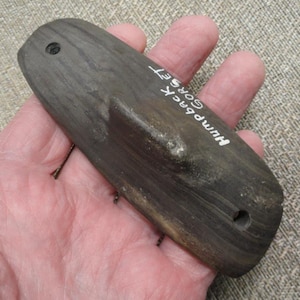 May include: A dark gray, elongated wooden object with a curved shape and a raised section. The text "Humpback Corset" is written on the surface. There are two holes drilled into the wood. The object is held in a hand.