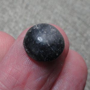 May include: A small, rounded, dark charcoal-gray stone with a smooth, polished surface and subtle, light-colored natural veining throughout. The object is circular in shape and features a slightly reflective, matte texture across its surface.