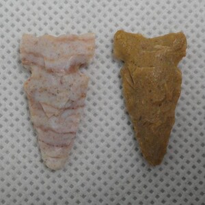 May include: Two stone arrowheads. One is a light pink with darker pink and white veining. The other is a solid tan color. Both have a similar shape, with a notched base and a pointed tip. The arrowheads are on a white background.