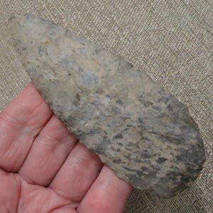 May include: A large, elongated, leaf-shaped stone tool with a pointed tip and a rounded base. The surface features a mottled, textured pattern in shades of gray, tan, and brown, created by flaked edges along the entire perimeter of the object.