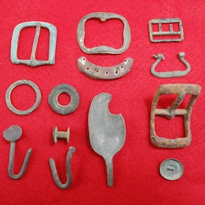 May include: A collection of antique metal hardware pieces, including buckles, rings, hooks, and a spoon-shaped object. The items are in various shapes and sizes, with a weathered, aged appearance. The background is a solid red.