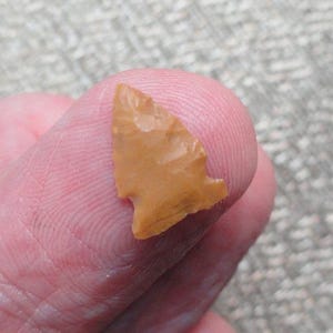 May include: A small, tan arrowhead with a triangular shape and a notched base. The arrowhead is made of stone and appears to be hand-chipped. The arrowhead is held between two fingers.