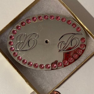 May include: A silver oval-shaped belt buckle with a pink rhinestone border. The buckle has two initials, "S" and "T", engraved in a cursive font.