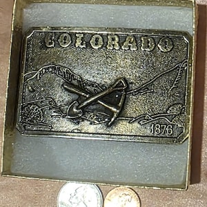 May include: A rectangular metal belt buckle with the word "COLORADO" and the year "1876" engraved on it. The buckle features a detailed landscape scene with crossed tools. The buckle is in a gold-tone box with two coins.