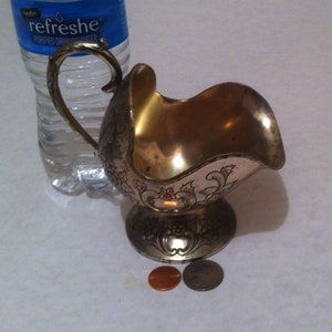 May include: A silver-colored metal creamer or sugar bowl with an ornate floral design. The bowl has a curved spout and a handle. The bowl is sitting on a white surface with two coins.