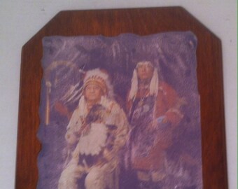 May include: A framed print depicting two individuals in Native American dress. The print is set within a dark wood frame with a decorative edge. The figures are wearing traditional clothing, including headdresses and ornate garments. The print has a vintage appearance.