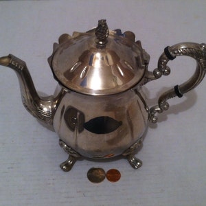 May include: Silver teapot with a decorative handle and three feet. The teapot has a lid with a finial on top.