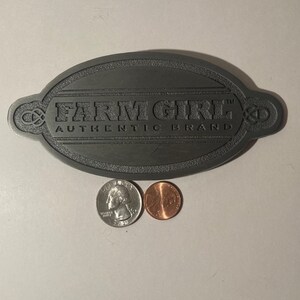 May include: A gray metal oval-shaped sign with the text "FARM GIRL™ AUTHENTIC BRAND" in black lettering.