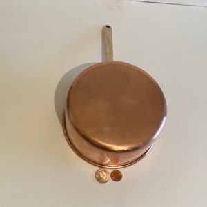以下が含まれることがあります： 木製の取っ手が付いた銅製の鍋。鍋は空で、白い表面の上に置かれています。