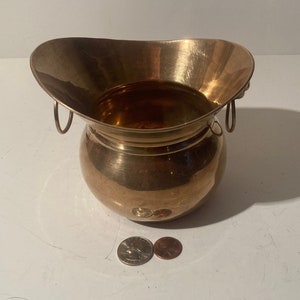 May include: A small, round, brass planter with two handles. The planter has a wide, flared top and a rounded bottom. It is sitting on a white surface.