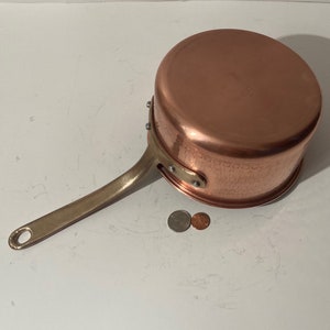 May include: A copper saucepan with a hammered finish and a brass handle. The saucepan is sitting on a white surface.