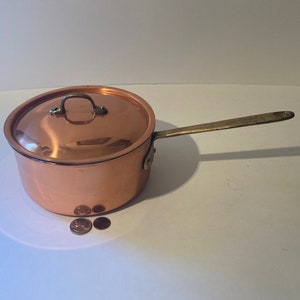 Op de afbeelding: Een koperen pan met een deksel en een messing handvat. De pan staat op een wit oppervlak.