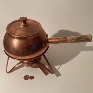 Könnte beinhalten: Kupfer-Fonduetopf mit Holzgriff und Ständer. Der Topf hat einen Deckel und steht auf einer weißen Oberfläche.