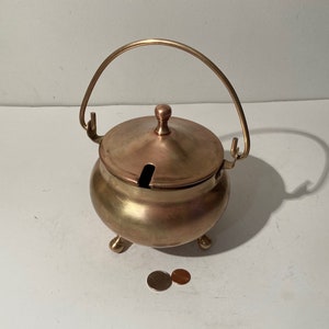 May include: A small, antique brass pot with a lid and a handle. The pot has three feet and a spout. The pot is sitting on a white surface.