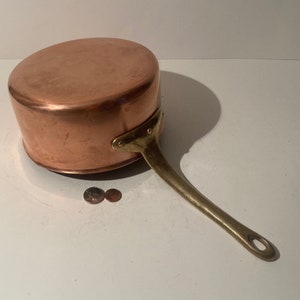 May include: Copper saucepan with a brass handle. The saucepan is sitting on a white surface with two copper pennies next to it.