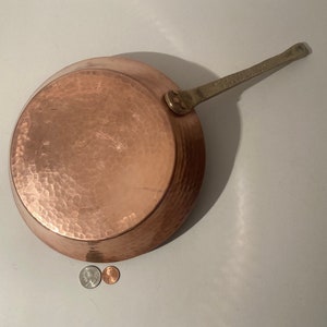 May include: A copper-colored, hammered-finish saucepan with a long, brass handle. The pan is round with a slightly sloped side. The handle is attached with a brass rivet. The pan is shown on a white surface with coins.