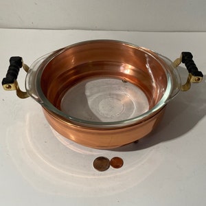 May include: A copper and glass serving dish with black handles. The dish has a copper base and a glass insert. The handles are made of black plastic and are attached to the dish with brass accents.