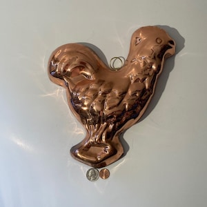 Puede incluir: Un molde de cobre en forma de gallo para hornear. El molde es brillante y tiene un diseño detallado. Es del tamaño de un plato de cena.