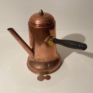 May include: Copper coffee pot with a black wooden handle and a brass knob on the lid. The pot is sitting on a white surface.