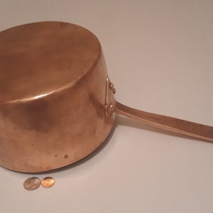 May include: A copper saucepan with a long handle. The saucepan is sitting on a white surface with two copper pennies next to it.