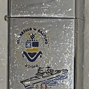 以下が含まれることがあります： 詳細なエンブレムが付いた銀色のライター。「U.S.S. ARTHUR W. RADFORD DD-968」の文字と船のイラストが特徴です。ライターは光沢のある反射面を持っています。エンブレムには円形のデザインも含まれています。