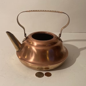 Puede incluir: Una tetera de cobre con asa y pico de latón. La tetera está sobre una superficie blanca.