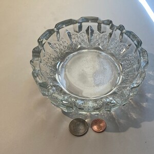 May include: A clear glass ashtray with a textured, geometric design. The ashtray is round and has a slightly raised rim.