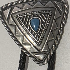 Op de afbeelding: Een zilveren bolo stropdas met een driehoekige hanger. De hanger heeft een ingewikkeld geometrisch ontwerp met een centrale turquoisekleurige steen. De randen zijn touwachtig en het koord is zwart.