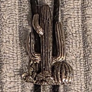 May include: A black leather bolo tie with a silver-colored cactus pendant. The pendant has detailed cactus features. The tie ends with silver-colored metal tips and is shown with a quarter and a penny.