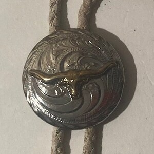 May include: A silver bolo tie with a braided leather cord and a round silver pendant featuring an engraved design of a steer head with gold horns.