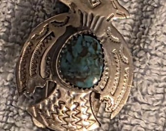 May include: A silver bolo tie with a turquoise stone center. The silver pendant features an engraved design and is attached to a black braided leather cord. A quarter and a penny are in the image.