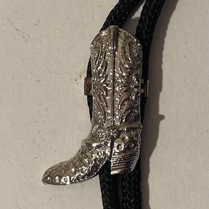 Puede incluir: Un corbatín bolo negro con un dije de bota de plata. La bota está detallada con un diseño occidental.