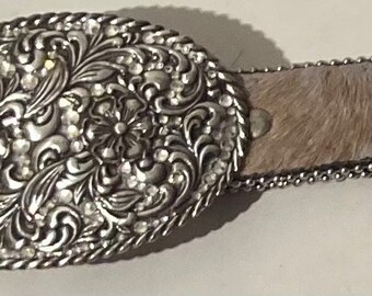 May include: A silver belt buckle with a floral design and rhinestones. The belt is made of brown leather with a white pattern and silver studs.