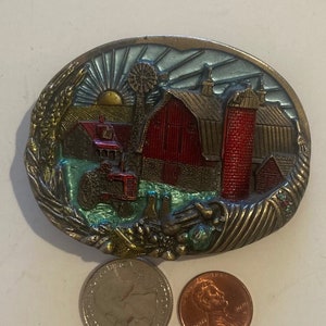 Może przedstawiać: Vintage metalowa klamra do paska z kolorowym emaliowanym wzorem czerwonego stodoły, traktora i wiatraka. Klamra jest otoczona złotą ramką z zielonym i niebieskim emaliowanym tłem.