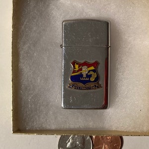 Könnte beinhalten: Ein silberfarbenes Feuerzeug mit einem farbenfrohen Emblem mit einem Schild, einem Gebäude und dem Text "U.S. MARINE CORPS". Das Feuerzeug befindet sich in einer kleinen, quadratischen Schachtel mit weißer Polsterung, neben einem Vierteldollar und einem Cent.